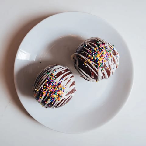 Close-up view of two dark chocolate spheres drizzled with white chocolate and sprinkles, sitting on a rustic wooden surface for a festive dessert presentation.  